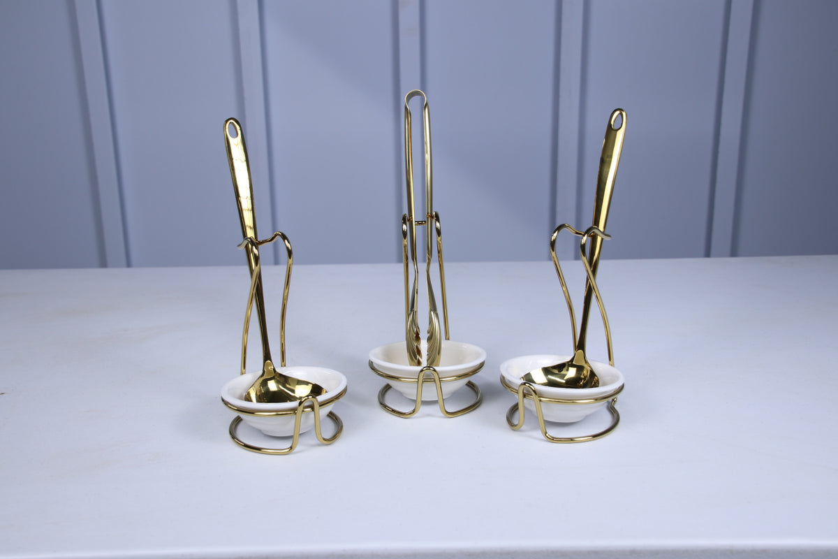Gold serving spoon, tongs and spoon in white ceramic holders, seen from the front