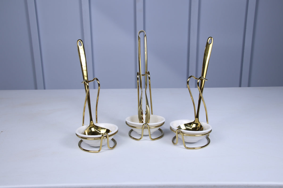 Gold serving spoon, tongs and second spoon, each resting in a white ceramic holder with a gold stand