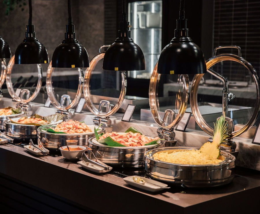 Stock photo: hotel buffet line with a row of round chafing dishes under black heat lamps, filled with hot dishes