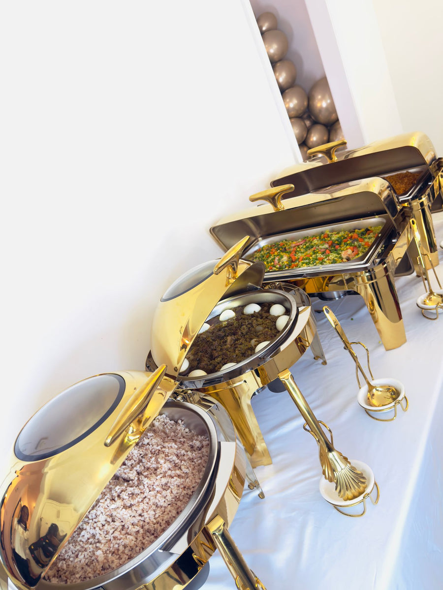Gold round and rectangle roll-top chafing dishes on a white buffet table at a customer's event, filled with rice, stew with boiled eggs and vegetable fried rice, with gold tongs and a ladle in holders